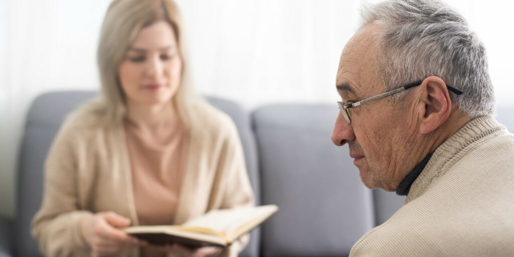 an elderly man sitting across from a woman holding a book